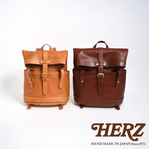 職人手作皮革雙口袋折蓋後背包，左為淺棕色，右為深棕色。兩款背包皆為皮質，設有雙前口袋、折蓋設計，並以皮帶及金屬扣環固定。