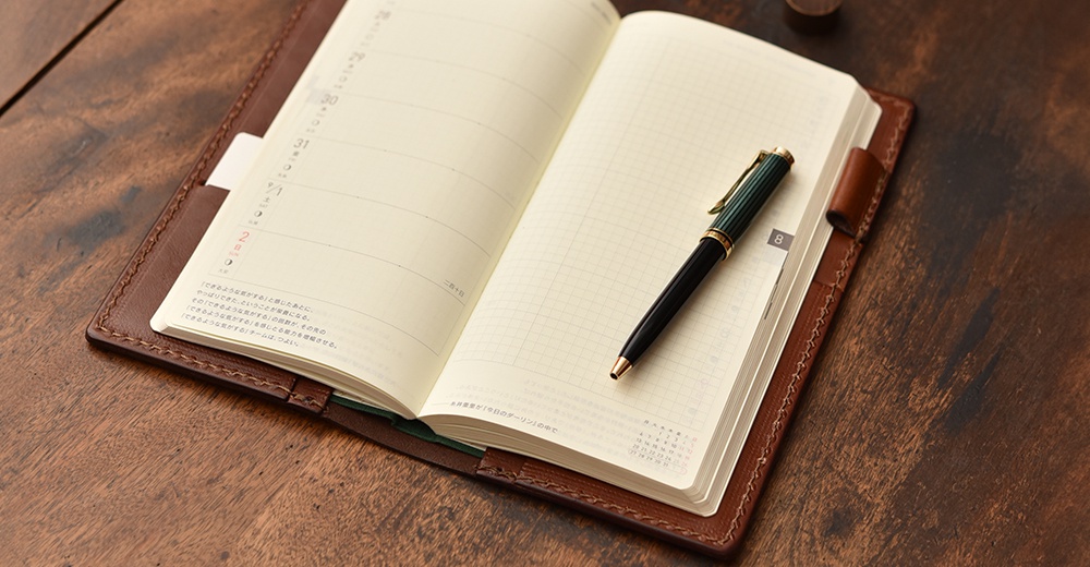 職人手作皮革旅行記事本書套，棕色皮革，內頁為日曆與方格紙，搭配一支綠色與金色的鋼筆。