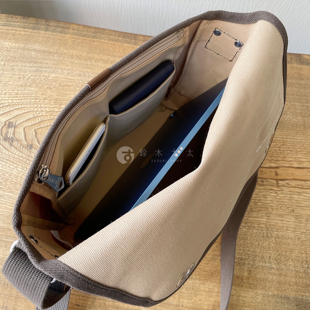 犬印純綿帆布雙扣郵差包內部，可見手機、皮夾收納隔層，以及一個薄型電腦。包包內裡為淺卡其色，外緣及背帶為深棕色。