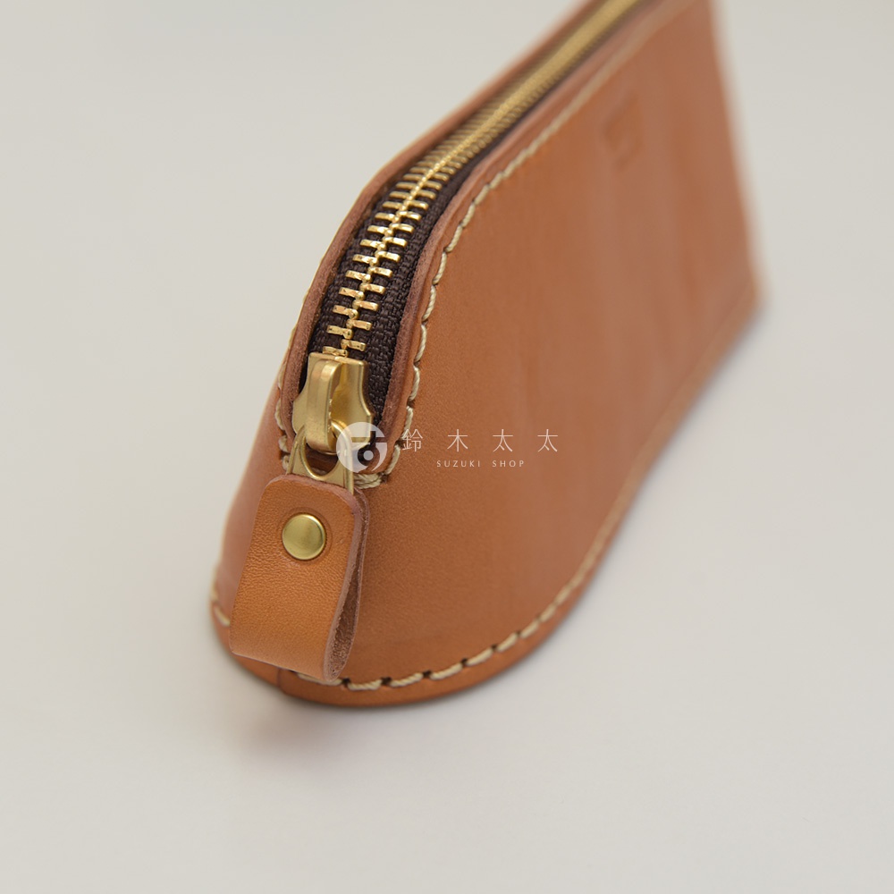 職人手作皮革三角弧形收納筆袋，淺棕色皮革，配有黃銅色拉鍊，縫線清晰可見，一側有皮革拉環和按扣。