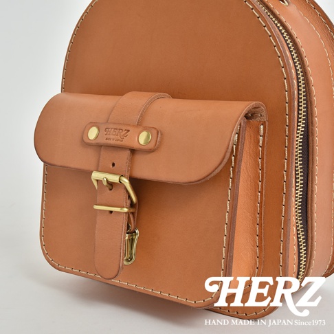 職人手作皮革迷你後背包，採用天然皮革製成，呈現溫潤的棕褐色。包身設計簡潔，配有翻蓋式前袋，以銅釦及皮帶扣固定，側邊設有拉鍊，展現細膩的手工縫線與高品質的五金配件。