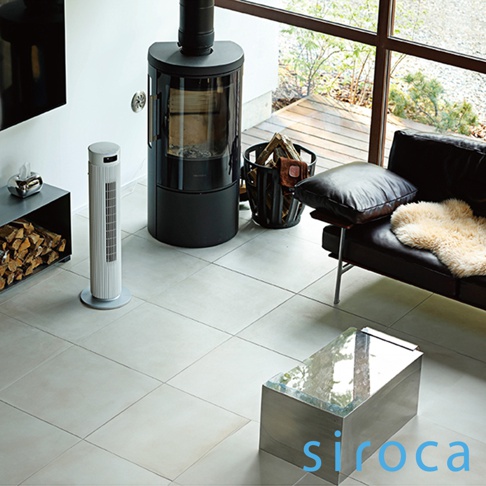 白色直立式循環扇，位於客廳地板上，旁邊有黑色爐具、柴火籃和黑色皮沙發。