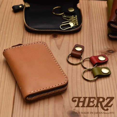 職人手作皮革多用途收納鑰匙包，主體為淺棕色皮革，配有金色拉鍊。旁邊有三個小鑰匙圈，分別為棕色、紅色和綠色皮革。上方展示一個打開的黑色皮革鑰匙包，內有四個金色鑰匙鉤。