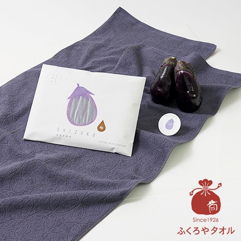 茄子紫色的薄毛巾，表面有著精緻的環狀紋理，旁邊擺放著兩顆新鮮茄子和印有茄子圖案的包裝紙。