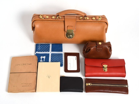 皮革製成的橫長形杜勒斯單肩包，搭配金屬鎖扣及提把。周圍擺放著棕色小零錢包、紅色長夾、棕色拉鍊皮夾、黑色拉鍊皮夾、深棕色卡夾、藍色白點圖案手帕，以及兩本不同顏色的筆記本。