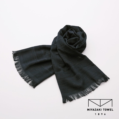 一條深色（接近炭黑色）的今治匹馬棉圍巾，材質輕盈，織紋細緻，兩端帶有流蘇，圍巾的一角捲起，整體呈現簡約優雅的風格。