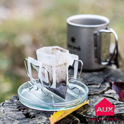 銀色金屬材質的可調式耳掛咖啡架，架上置有滴漏式咖啡濾袋，濾袋內有咖啡粉，後方有一個銀色金屬咖啡杯。