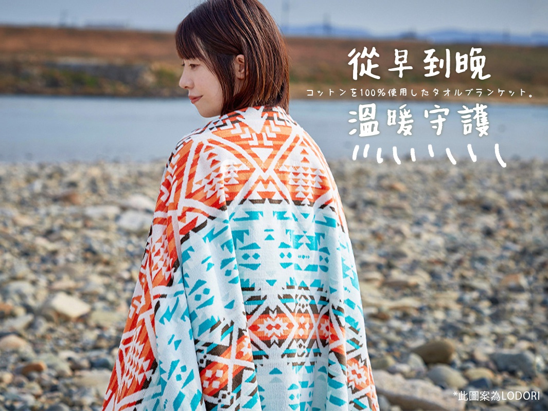 一位女性背對鏡頭，身上披著一件異國民族風萬用毛巾毯，毛巾毯以橘色、藍色、白色和黑色交織出幾何圖案，材質看起來柔軟。