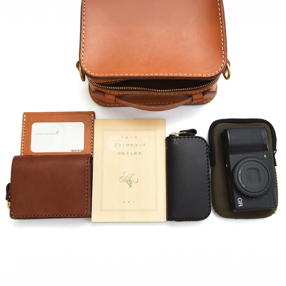日本HERZ職人手作皮革迷你小方包，旁邊擺放著一個皮夾、一本日文書以及一台相機。