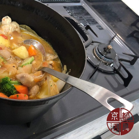 不鏽鋼萬用料理大勺，長柄設計，勺頭部分正在盛取鍋中燉煮的雞肉、馬鈴薯、紅蘿蔔和花椰菜等燉菜，勺柄上有一個圓形孔洞，置於黑色燉鍋內，鍋旁是廚房爐灶。