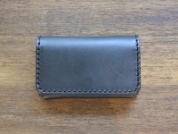 深灰色職人手作皮革Organ名片夾，表面光滑，邊緣以對比色縫線手工縫製，呈長方形，置於木質紋理背景上。