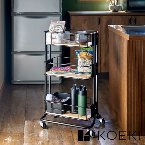 黑色三層置物推車，帶有木質層板，上方層架有零食包、罐子和紙盒，中間層架有零食包和毛巾，下方層架有黑色收納盒和兩個水壺，推車配有滾輪。