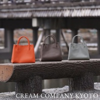 三款皮革手提托特包，分別為亮橙色、深棕色和淺灰色，皆配有提把，擺放在木質長凳上。