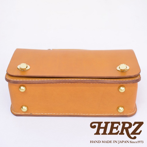 職人手作皮革經典兩用雙扣迷你單肩包，採用天然皮革製成，呈現溫潤的焦糖棕色。包款設有兩個金色旋轉鎖扣，側邊亦有兩個金色鉚釘裝飾，縫線細緻。