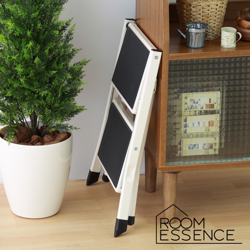 米白色折疊多用樓梯椅，兩層踏板，黑色防滑表面，椅腳有黑色保護套，倚靠在木紋櫃旁，旁邊有一盆綠植。