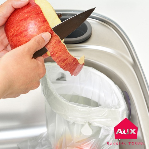 一個人在水槽上方削蘋果，蘋果皮被丟進一個掛在水槽邊緣的白色廚餘袋架裡，架上附有 AUX 品牌標誌。