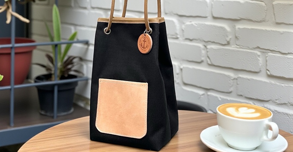 一個黑色束口帆布包，配有棕褐色皮革口袋和船錨吊飾，旁邊放著一杯裝飾著拉花的咖啡。