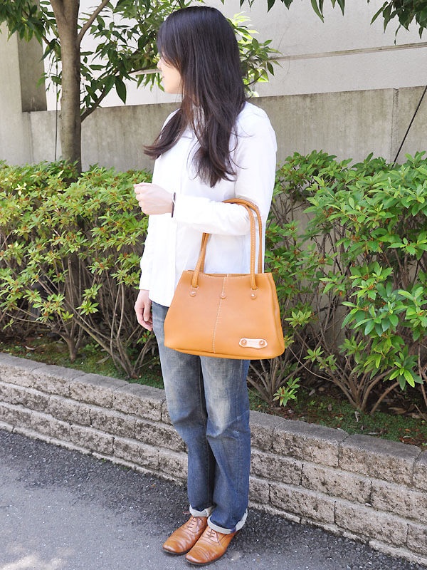 焦糖色皮革手提托特包，設有兩個提把和一個皮革裝飾片，包身線條簡潔，搭配藍色牛仔褲和棕色皮鞋。