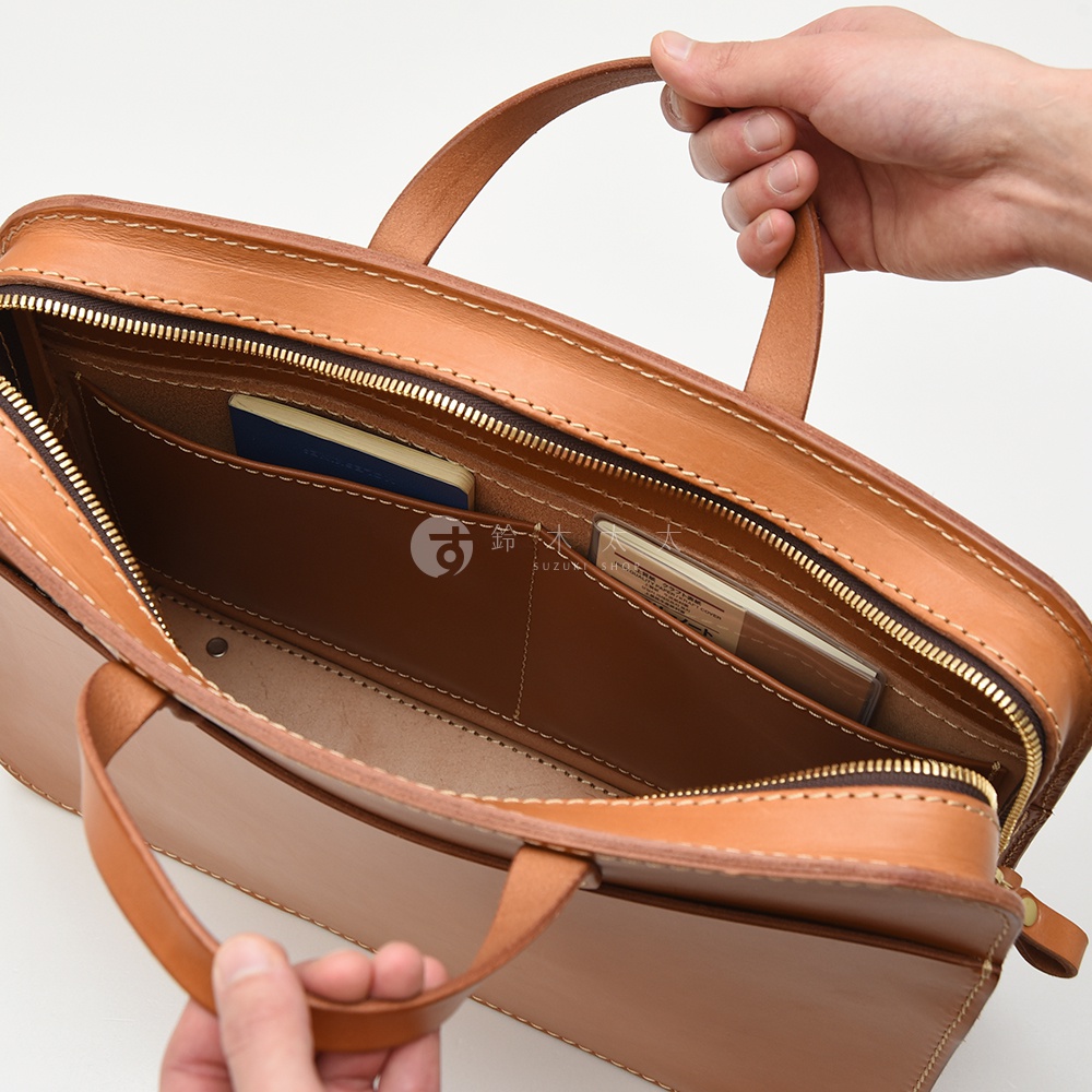 職人手作皮革兩用商務公事包，呈現溫潤的淺棕色調，以真皮材質打造，細膩的縫線展現高品質工藝。包內設有多個隔層，可收納護照、卡片等小物，展現其功能性與質感。