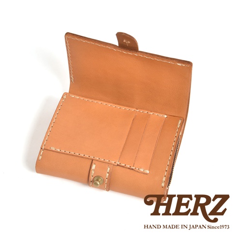 這是一張日本HERZ品牌職人手作皮革中型皮夾的產品照，採用原色植鞣皮革製成，表面具有溫潤的棕色質感。皮夾呈現打開的視角，展現出內部精細的縫線工藝，設有多個卡夾空間與金屬按扣設計，整體風格簡約復古，右下角印有HERZ品牌商標與創立年份。