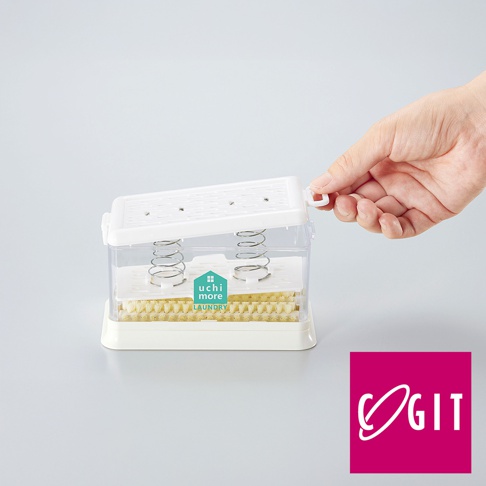 白色塑膠製的免沾手泡沫洗衣皂盒刷，透明盒內有兩組彈簧及黃色刷毛，上方為白色掀蓋，一隻手正將蓋子打開。