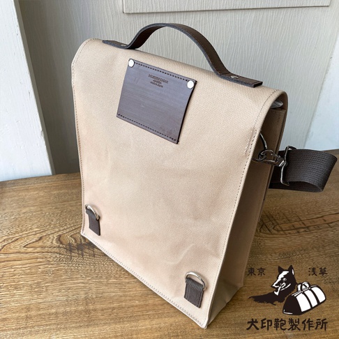 米色純棉帆布製成，帶有皮革提把及側肩背帶，正面有深棕色皮革方形裝飾片，並印有品牌標誌。