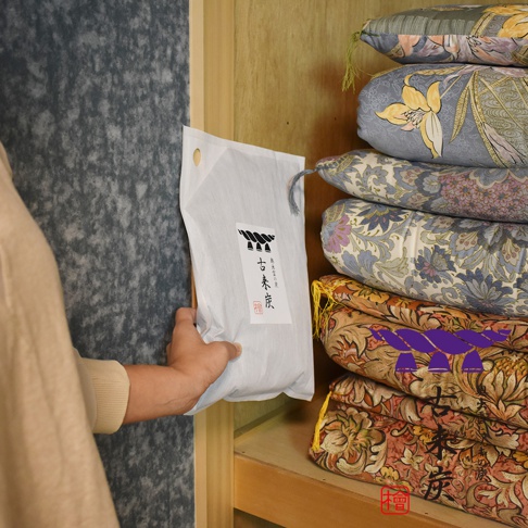 一隻手拿著印有「古來炭」字樣的白色包裝袋，袋上印有日式圖案。旁邊的木架上堆疊著數個顏色豐富、印有花卉圖案的布製抱枕。