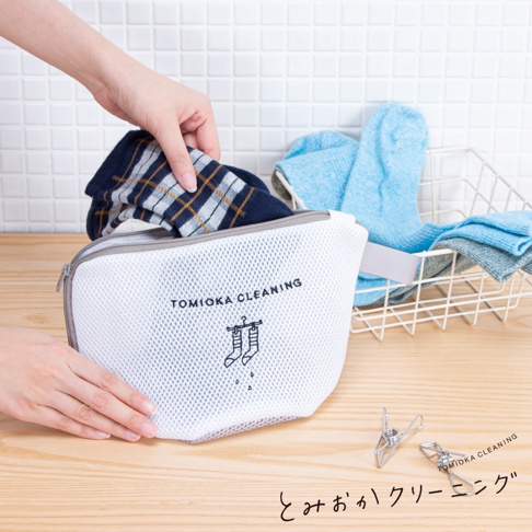 白色網狀貼身衣物洗衣袋，有灰色拉鍊和提帶，印有「TOMIOKA CLEANING」字樣及襪子圖案。一隻手正將一雙藍色格紋襪子放入袋中，背景有籃子裝著藍色和灰色襪子。