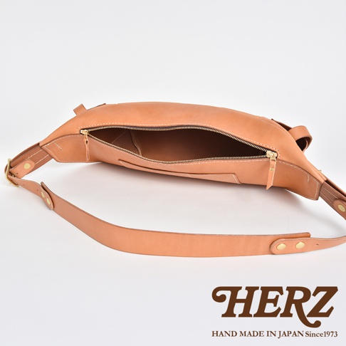 HERZ 職人手作斜背包，使用Stare皮革，展現自然色澤，拉鍊開啟露出內部，搭配品牌標誌和產地資訊