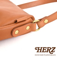 HERZ 職人手作皮革半圓馬鞍包 CK-17 細節，展示焦糖色皮革製作的包款，金屬旋鈕及 D 型環五金配件，上方印有 HERZ 品牌標誌，下方標示 HAND MADE IN JAPAN Since1973。