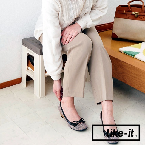 奶油色與灰色塑膠製成的玄關穿鞋收納椅，帶有兩層鞋架空間，可收納一雙鞋子。一位女士穿著米色長褲和格子平底鞋，坐在椅子上。