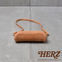 一個淺棕色皮革製成的長方形斜背包，帶有可調節的細長背帶，背包表面呈現細膩的紋理，置於水泥地面上，右下角有HERZ品牌標誌與HAND MADE IN JAPAN Since1973字樣