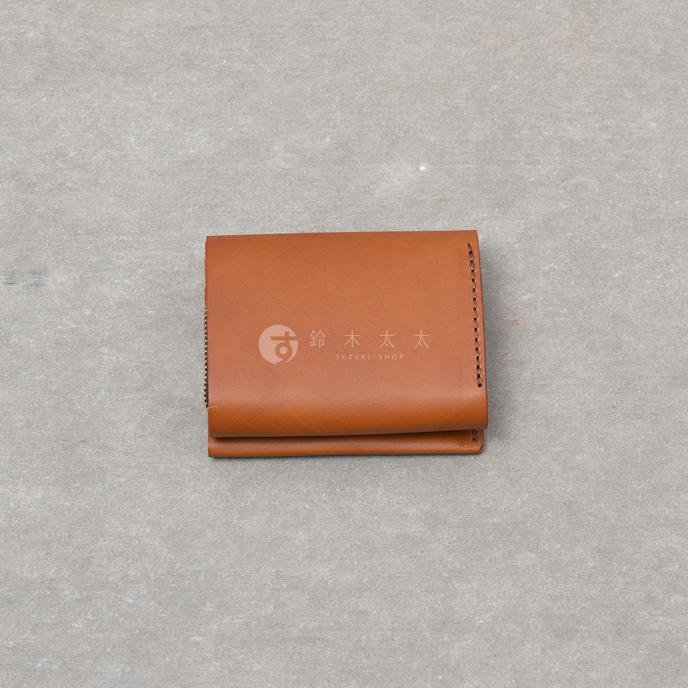 一個焦糖色皮革製成的雙折迷你短夾，表面印有鈴木太太的品牌標誌與文字，錢包右側邊緣有精緻的手縫線細節，設計簡約俐落，呈現出質感溫潤的義大利皮革紋理。