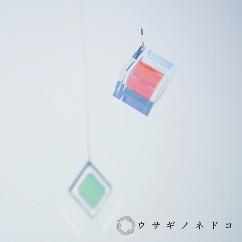 透明稜鏡立方體吊飾，內部有粉紅色層次，透過光線折射呈現藍紫色光澤。下方懸掛著一個綠色菱形吊飾。