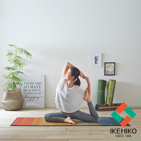 一位女性在色彩漸層的藺草榻榻米瑜珈墊上做著瑜珈動作。墊子為大地色系，搭配綠色及紅色交織的logo。背景有植物、裝飾畫及捲起的瑜珈墊。