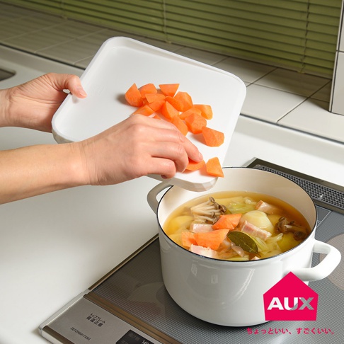 一隻手拿著白色迷你砧板，上面有切塊的胡蘿蔔，正準備將胡蘿蔔倒入裝有燉菜的白色鍋中。