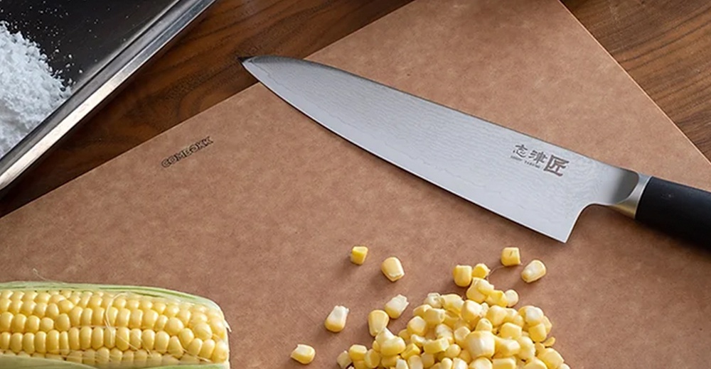 一把銀色不鏽鋼主廚刀，刀身上有明顯的紋路，黑色刀柄。刀旁有半根玉米和散落的玉米粒，以及一個金屬容器。