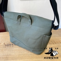 軍綠色純棉帆布雙扣郵差包，有提把和側邊口袋，搭配黑色肩帶，置於木質桌面上。