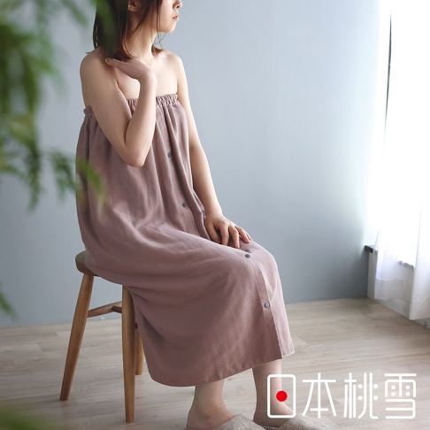 一位女性身穿粉色棉紗混紡平口鬆緊浴裙，浴裙上有暗色鈕扣設計，女性坐在木凳上，背景為淺灰色牆壁與白色窗簾。