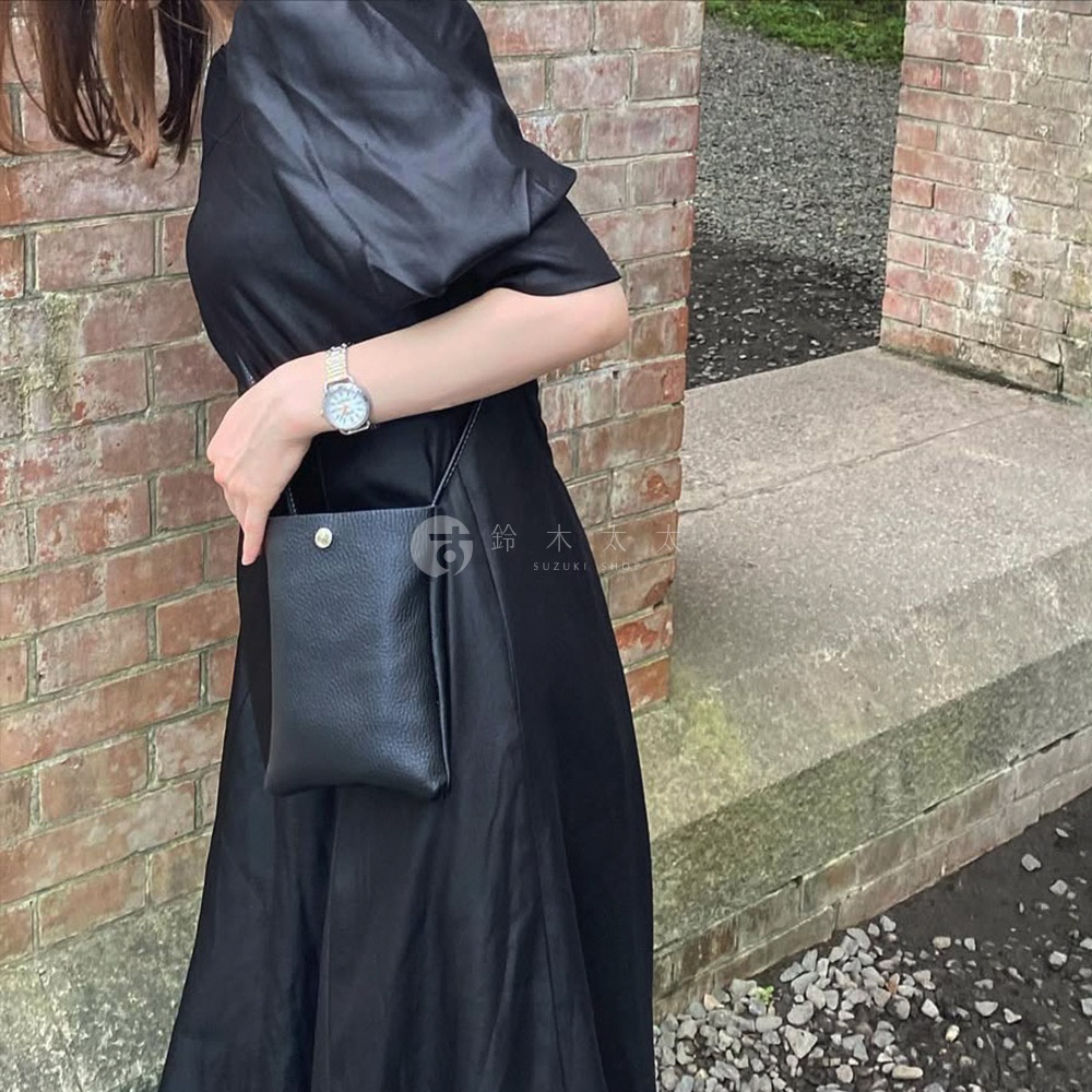 黑色皮革隨身手機包，附有金屬按扣和細長肩帶，由身著黑色洋裝的模特兒展示。