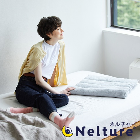 一位身穿黃色開襟衫、白色T恤和牛仔褲的女士，坐在床上，手觸碰著一個灰色的毛巾枕，枕頭上有多道捲曲紋理。