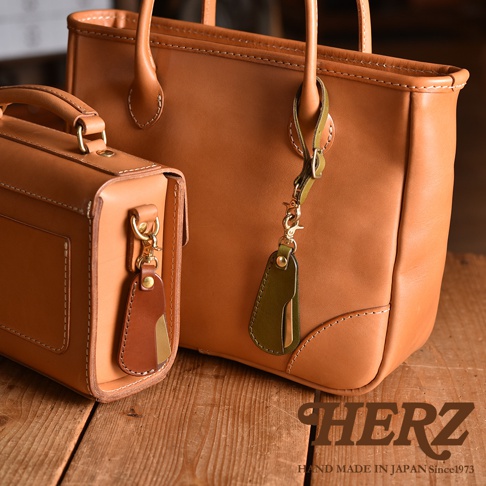 兩款TAN色系皮革包，其中一款為手提包，另一款為小方包，皆有附上金屬扣環及皮革吊飾，吊飾由棕色與綠色皮革及金屬片組成。