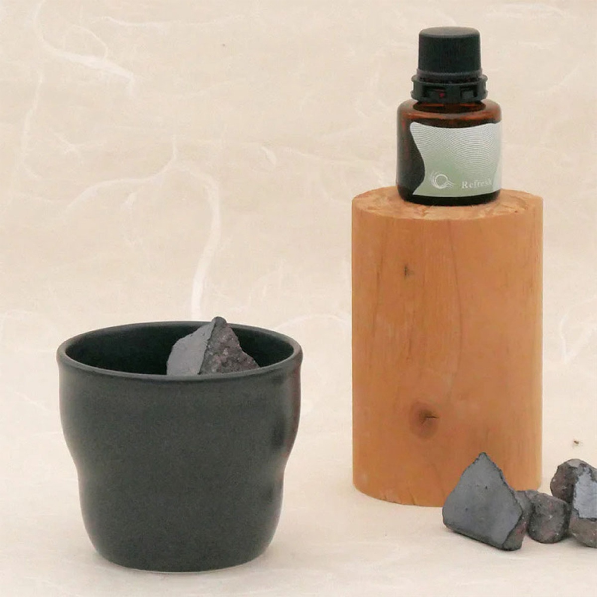 黑色陶瓷杯中放著幾塊深灰色的石頭，杯子旁邊散落著幾塊同樣的石頭，背景是一個木質圓柱體，上面放著一瓶帶有標籤的棕色精油瓶，標籤上寫著「Refresh」。