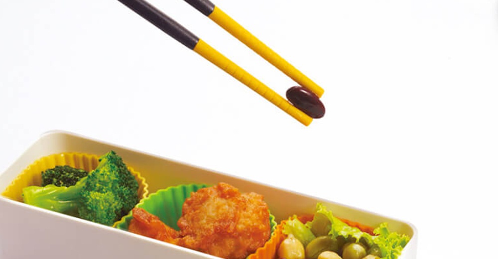 一雙黃色筷子夾著一顆深色豆子，上方背景為白色，下方是裝有食物的便當盒，食物包含花椰菜、炸物和沙拉。