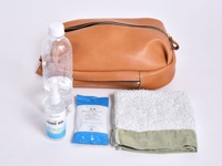 職人手作皮革立體運動斜背包 (F-9)，淺棕色牛皮材質，包身呈現圓潤立體造型，配有金色拉鍊。旁邊擺放一瓶礦泉水、一瓶酒精乾洗手液，以及一包濕紙巾和一條毛巾。