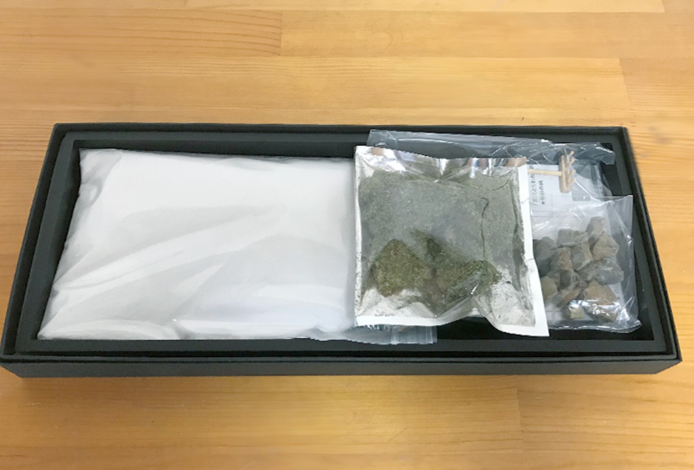 黑色長方形盒中，左側為白色粉末裝於透明塑膠袋，右側則有裝有深綠色苔蘚狀物體的銀色包裝袋，以及裝有小石塊的透明包裝袋。