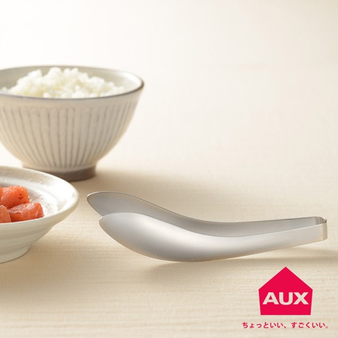 一雙銀色金屬不沾桌配料專用夾，夾身呈現流線型設計，置於淡黃色背景布上，旁邊有一個裝有紅色塊狀食物的白色小碗和一碗白米飯。