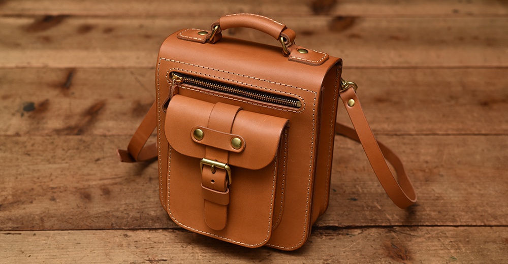 棕色皮革製成的手提/單肩兩用包，設有頂部提把、拉鍊開口主袋、翻蓋前袋及黃銅色金屬釦具。