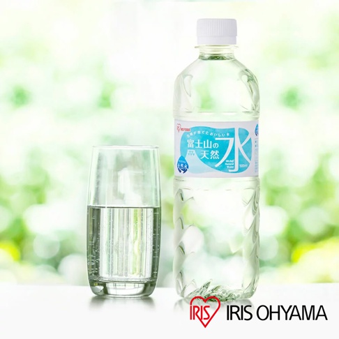 透明寶特瓶裝的富士山礦泉水，瓶身有藍色和白色標籤，標示「富士山の天然水」字樣。旁邊有一杯裝有水的透明玻璃杯。背景為綠色模糊的自然景觀。