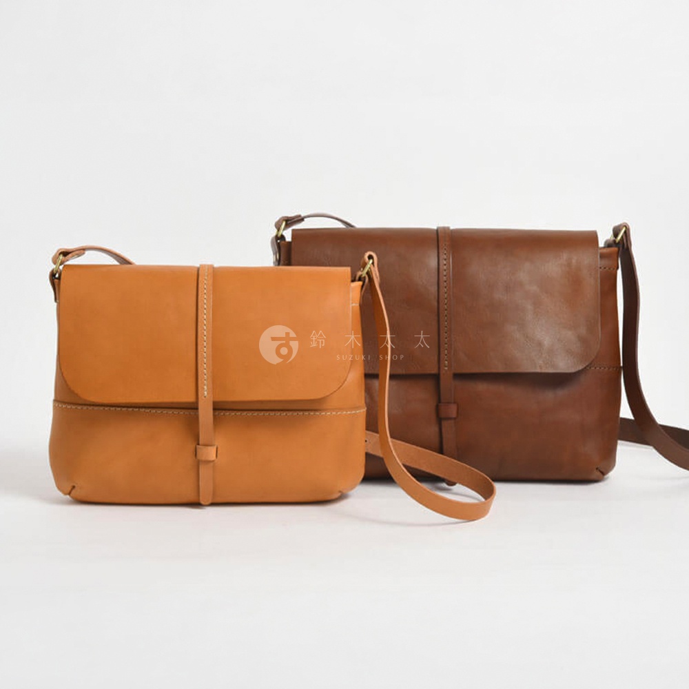 日本HERZ職人手作皮革細扣帶馬鞍包 CK-3，以茶色和棕色兩種顏色呈現，兩款包包以斜角對角線擺放，其中茶色包包置於前景，棕色包包置於後景，兩款包包均採用細緻的皮革材質，簡約的設計和實用的馬鞍包造型，凸顯職人手作的質感。